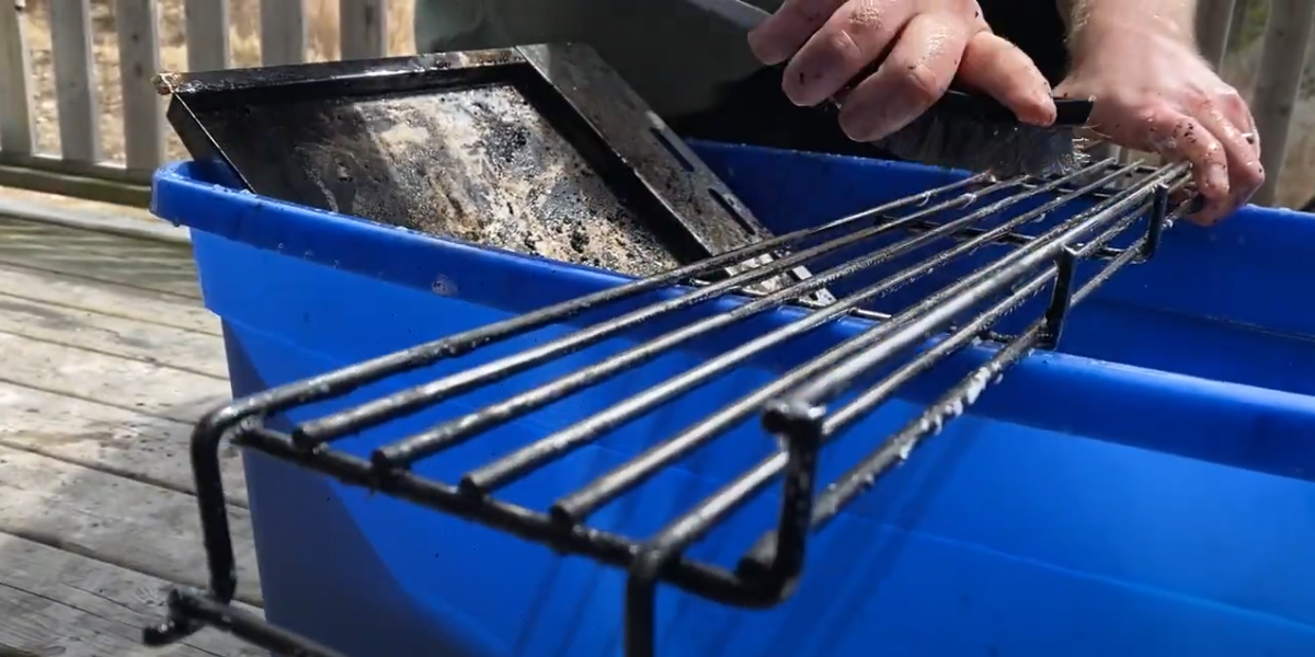 Grill grates and heat shields are cleaned in a blue bucket of soapy water with a wire bristle brush