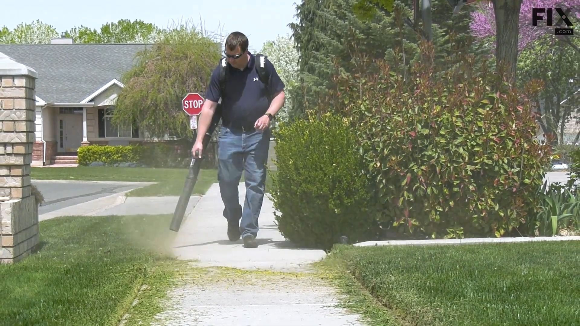 Man in a suburban neighborhood using a backpack leaf blower to clear grass clippings and debris from a sidewalk