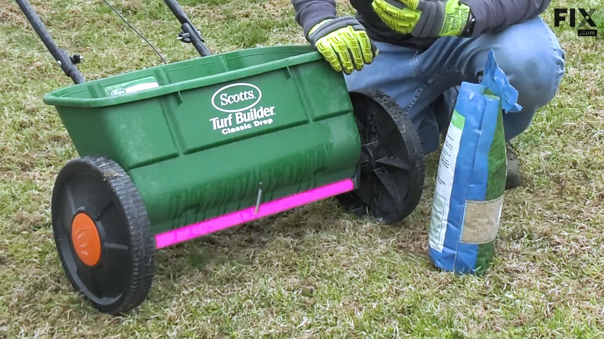 Man wearing protective gloves crouching between a bag of fertilizer and a drop fertilizer spreader with its bottom door highlighted in pink