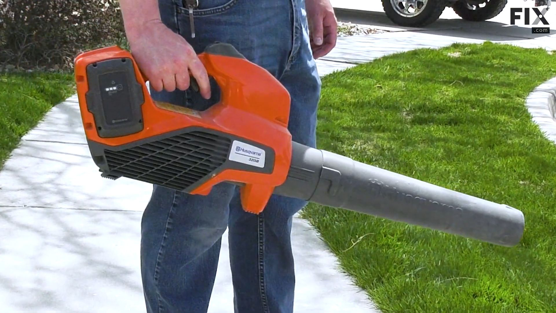 Man standing on a pavement in a suburban neighborhood while carrying a small, orange, handheld leaf blower model