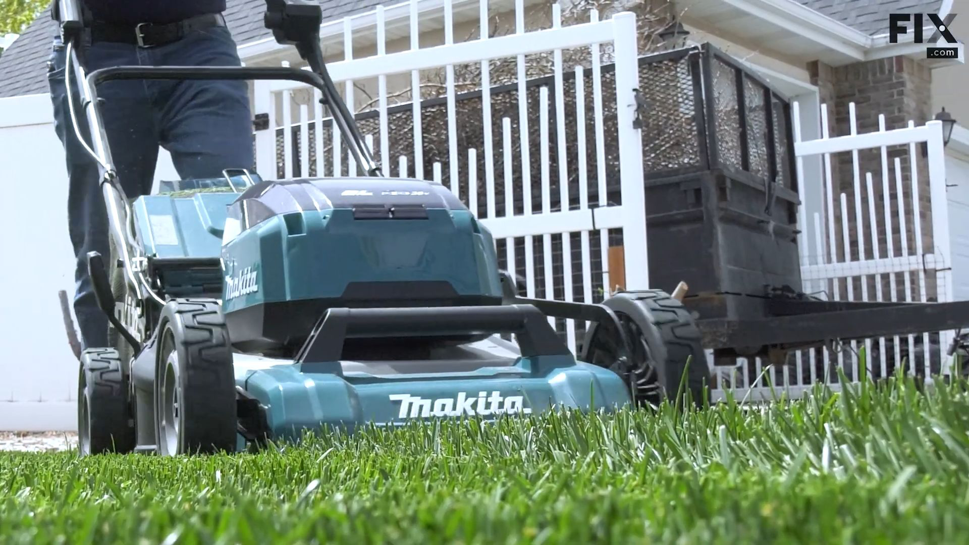 Man pushing a blue, battery-powered, self-propelled lawnmower through a front lawn of a suburban home with a white fence