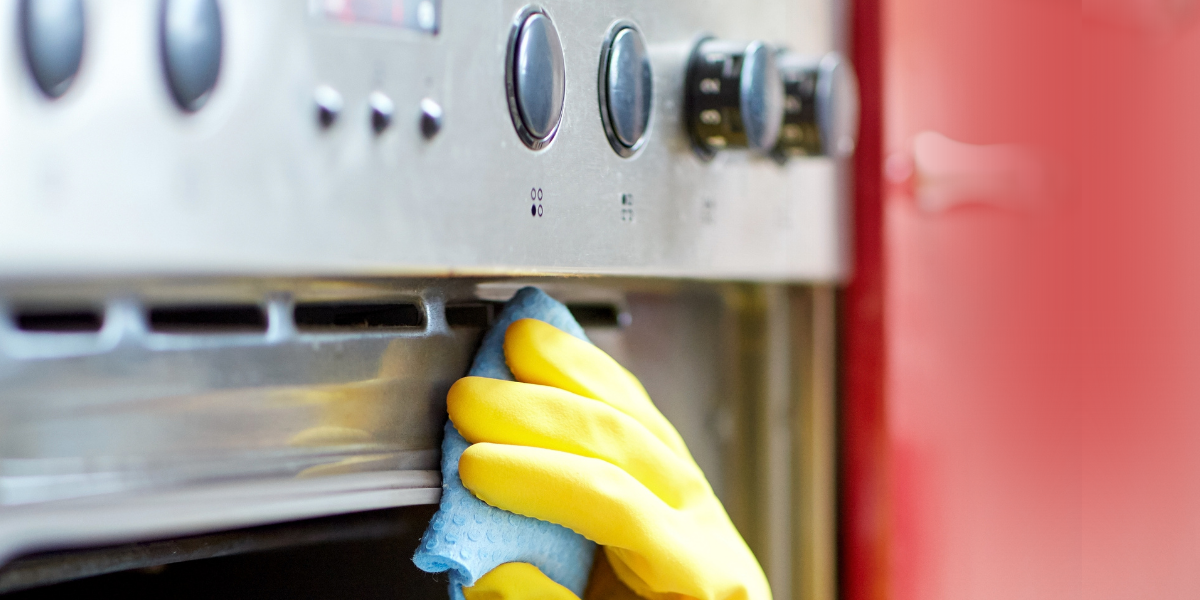 Hand in a thick yellow rubber glove wiping a stainless steel range oven frame with a soft blue cleaning sponge 