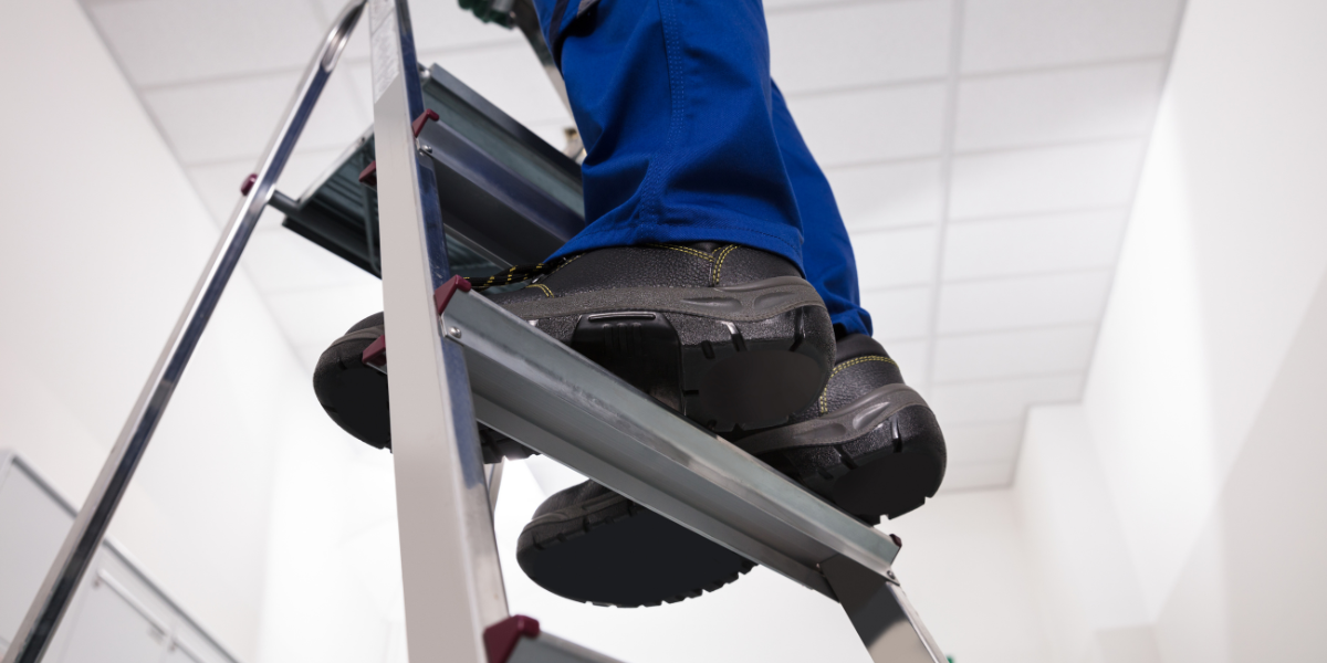 A person wearing work foots stands with both feet on a ladder rung. Ladder safety is an important part of DIY home repairs.