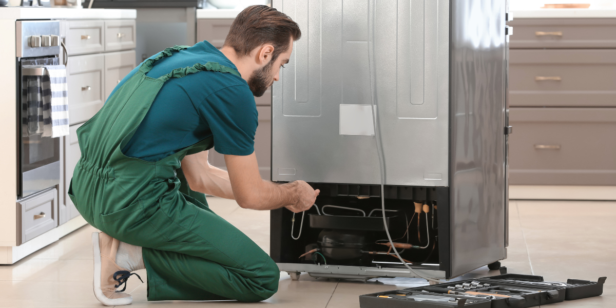 A repair technician kneeling on the kitchen floor, black toolbox open beside him, working on the lower back panel of a fridge