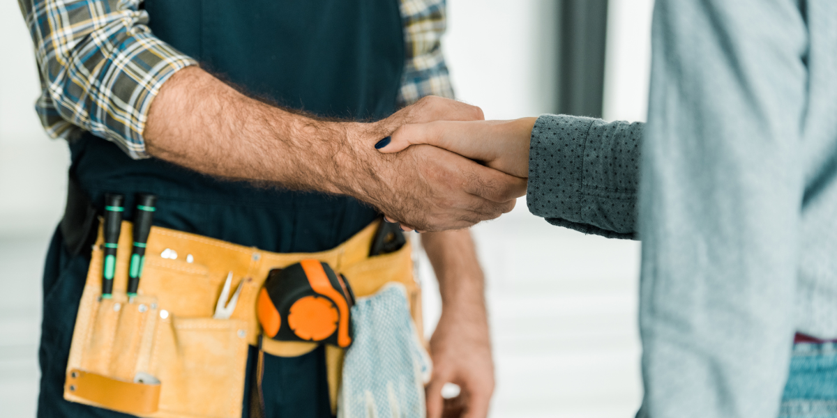 A repair technician, dressed in a flannel top and sporting a fully equipped yellow toolbelt, shakes hands with a customer