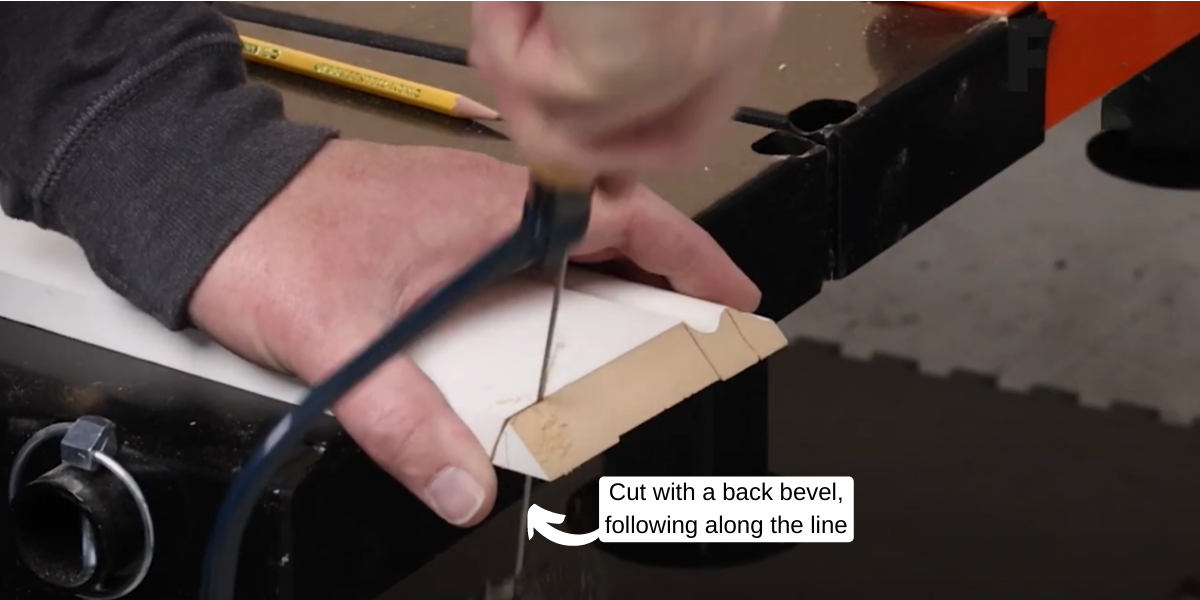 A hand holds a piece of baseboard trim while another hand holding a cope saw cuts into the trim using a back bevel technique. .