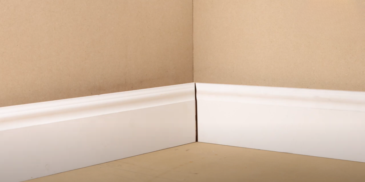 An inner corner of a wall shows two pieces of baseboard trim. There is a gap between the joint from the wood trim shrinking.