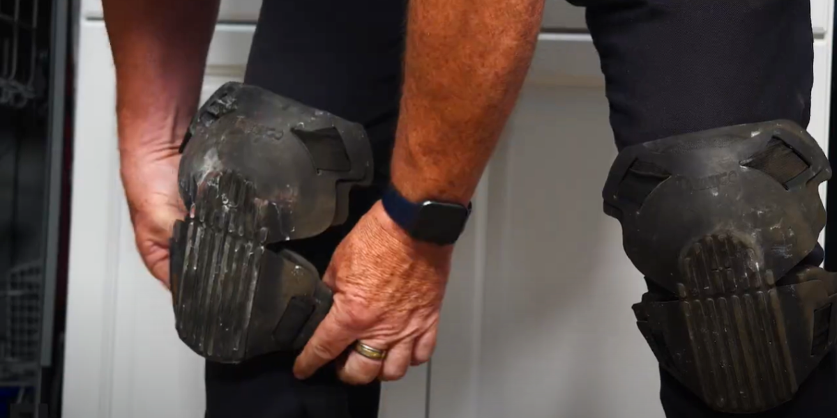 A man wearing a smartwatch is placing black kneepads over his right knee. The left knee is already covered by a kneepad.