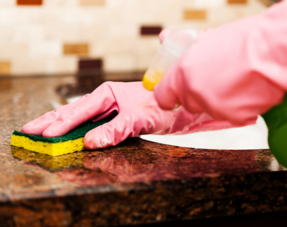 Repair technician cleaning the granite countertop of a kitchen sink with a sponge, a spray bottle and a pair of rubber gloves