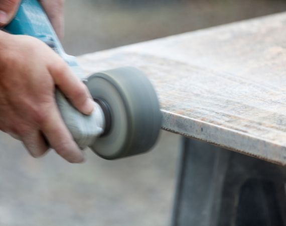 Repair technician smoothening out the edges of a slab of granite atop a worktable, with the help of a blue orbital polisher.